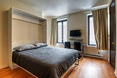 a bedroom with a bed and a desk and two windows at Studio Stays Downtown Montreal in Montréal