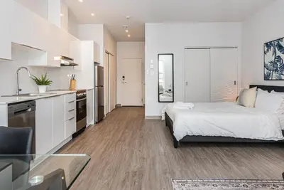 a white bedroom with a bed and a kitchen at Modern and New Studio in the Heart of the City by Den Stays in Montréal